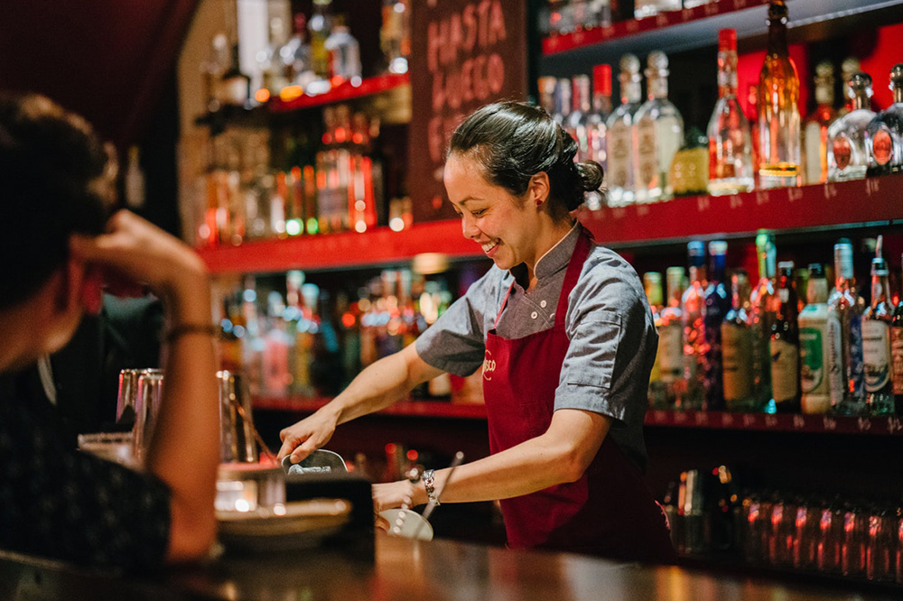 En leende bartender. Hon står och mixar drinkar till kunderna som sitter på andra sidan bardisken.