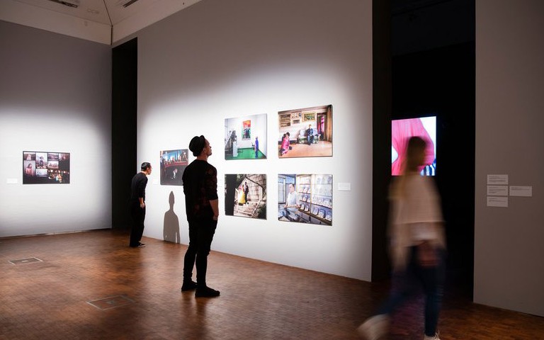 Ett konstgalleri, två personer kollar på färgglada tavlor som sitter på en vit vägg, en suddig gestalt går förbi