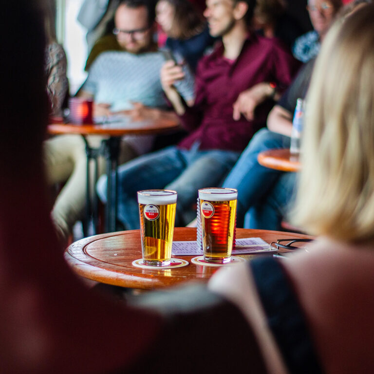I förgrunden ser vi två glas öl som står på ett bord och människors ryggar samt några som sitter vid bord.