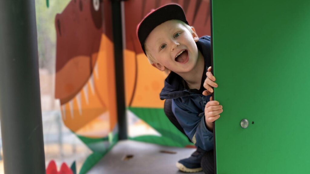 En pojke med keps sitter i en lekplats. Han sitter bakom en grön vägg. Pojken ser glad ut och skrattar.