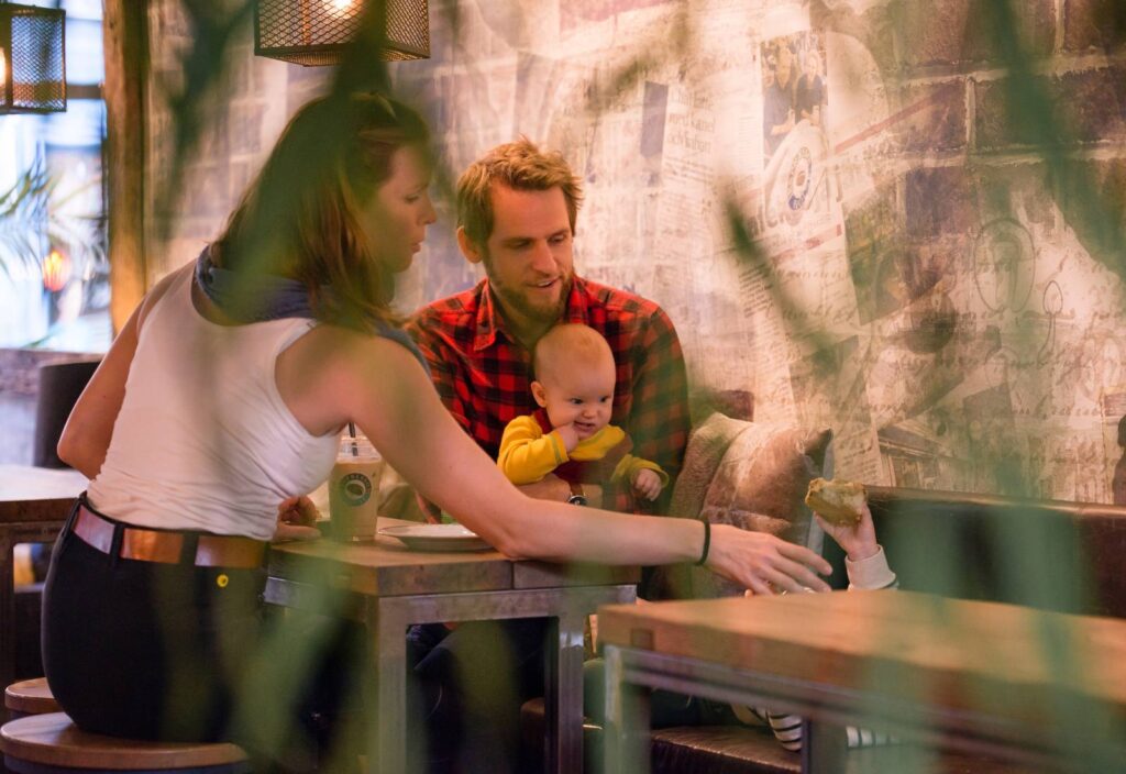 En familj sitter på ett café och fikar, ett litet barn sitter i knäet på en fadersgestalt.