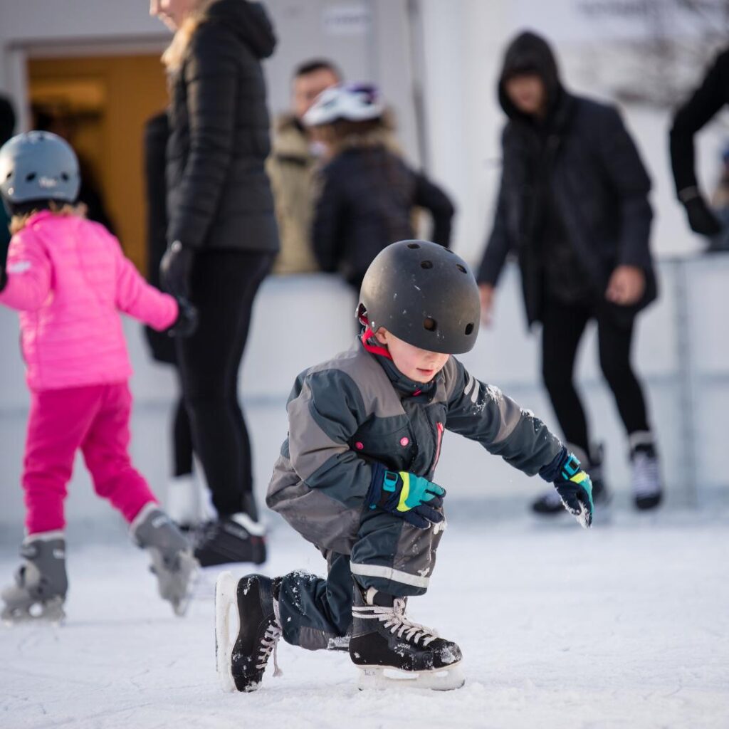 ett litet barn som har på sig overall och hjälm och åker skridsko på en utomhus isbana