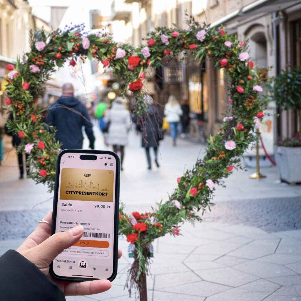 en bild på ett härta i väster och blommor, framför ser man en hand som håller i en mobil med en bild på ett digitalt citypresentkort i guldig färg