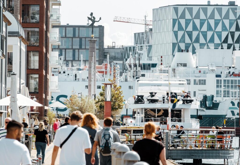 En bild som visar delar av norra hamnen i Helsingborg. MAn ser människor bakifrån som går på gatan, solen skiner, ett stort fartyg i bakgrunden och uteserveringar.