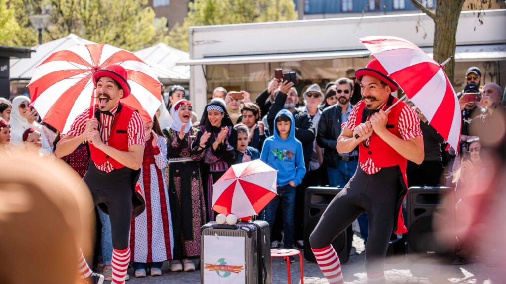 Två clowner iklädda röda hattar och röda och svarta kläder dansar med vit- och rödrandiga paraplyer. Bakom dom står en publik.
