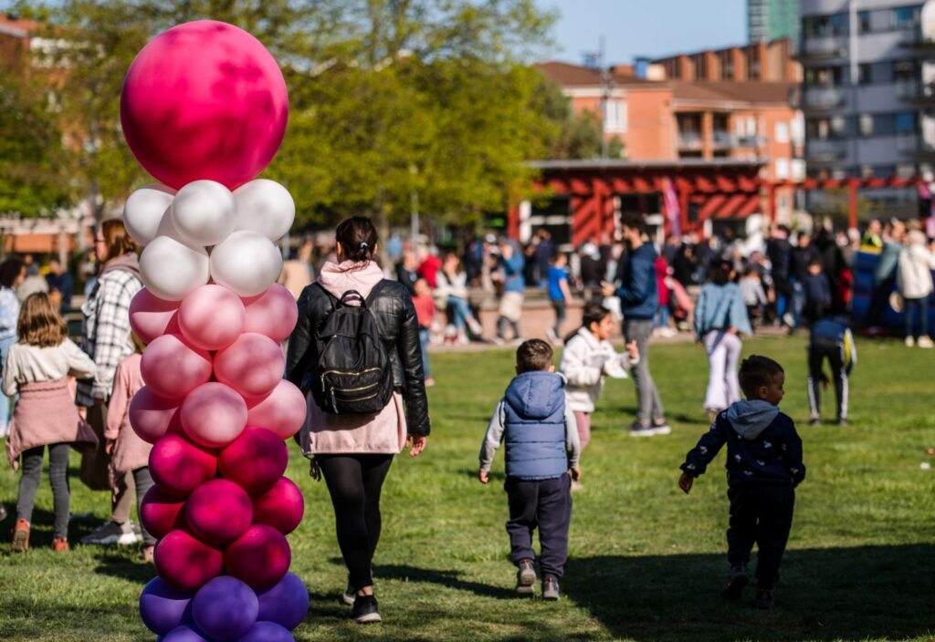 Utomhus i en park, i förgrunden ser man ballonger i rosa, vitt och blått. Många barn och vuxna går på gräsmattan.