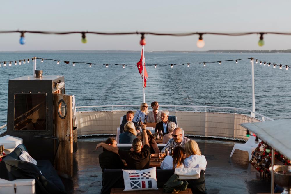 Aktern på en båt. På båten sitter folk runt bord och dricker drinkar. Över borden hänger lampor i olika färger. I bakgrunden ser man sundet mellan Helsingborg och Helsingör.