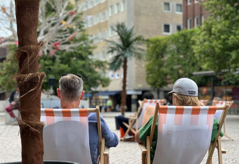 Två personer som sitter i solstorlar med ryggen till och tittar ut över ett torg med palmer och livemusik