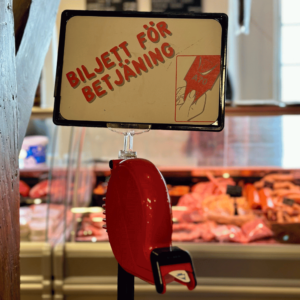 En röd biljettmaskin för kölappar med en en vit skylt med röd text ovanför. Det står Biljett för betjäning på skylten. I bakgrunden ser man en disk med kött och chark.