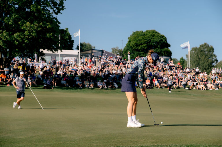 Golfbana, en kvinna i förgrunden som ska slå klubban, i bakgrunden syns massor av människor som tittar på.