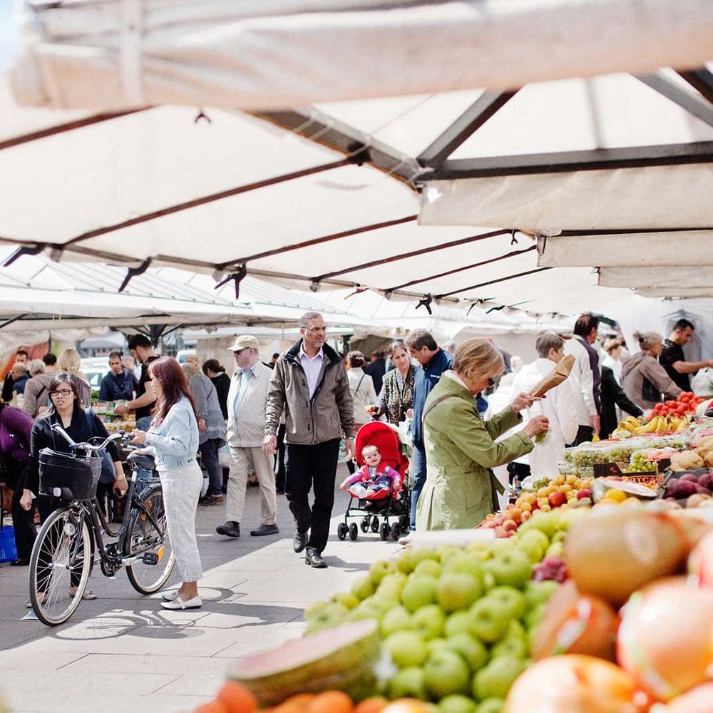 Marknaden på Gustav Adolfs torg. Marknadsstånd med frukt i förgrunden och i bakgrunden ser man människor som går och handlar och tittar på varorna.