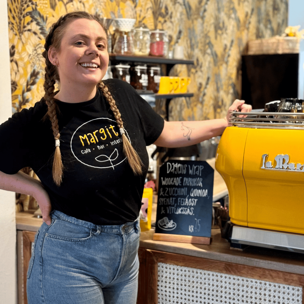 En kvinna i flätor står bredvid en gul kaffemaskin. Hon har blåa jeans och en svart tröja där det står Margit Café på. I bakgrunden ser man en vägg med gul tapet med mönster och en hylla med kaffekoppar.