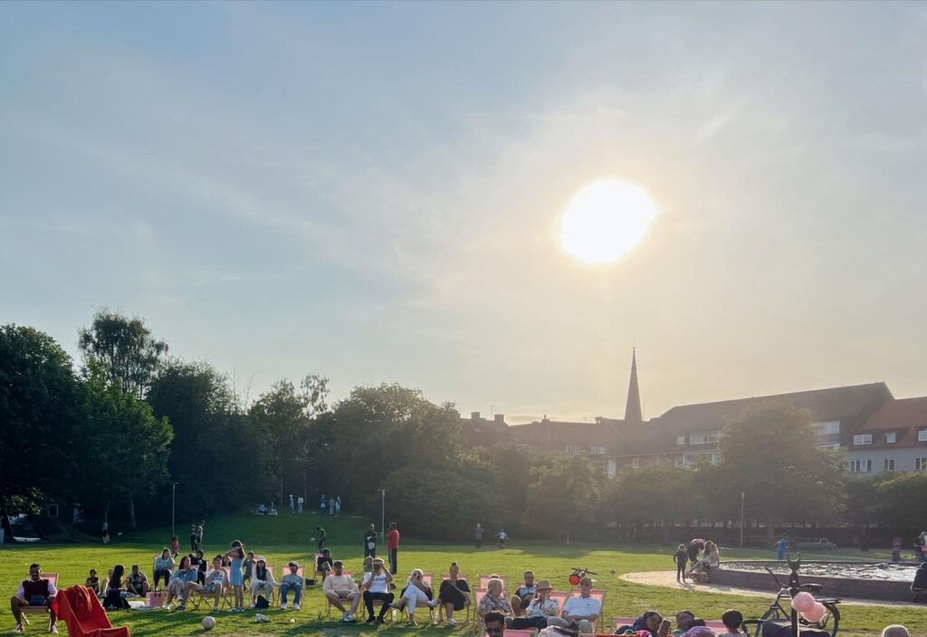 Utomhus i en park men solen som lyser skarp, det sitter folk på solstolar och tittar på en konsert