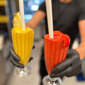 Två händer med svarta plasthandskar håller i två glas med smoothie. Den vänstra är gul med en ananas i och den högra är röd med en jordgubbe i.