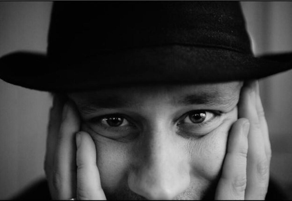 Ett svartvitt foto med en närbild på en kille i svart hatt, man ser endast ansiktet och han håller händerna på kinderna