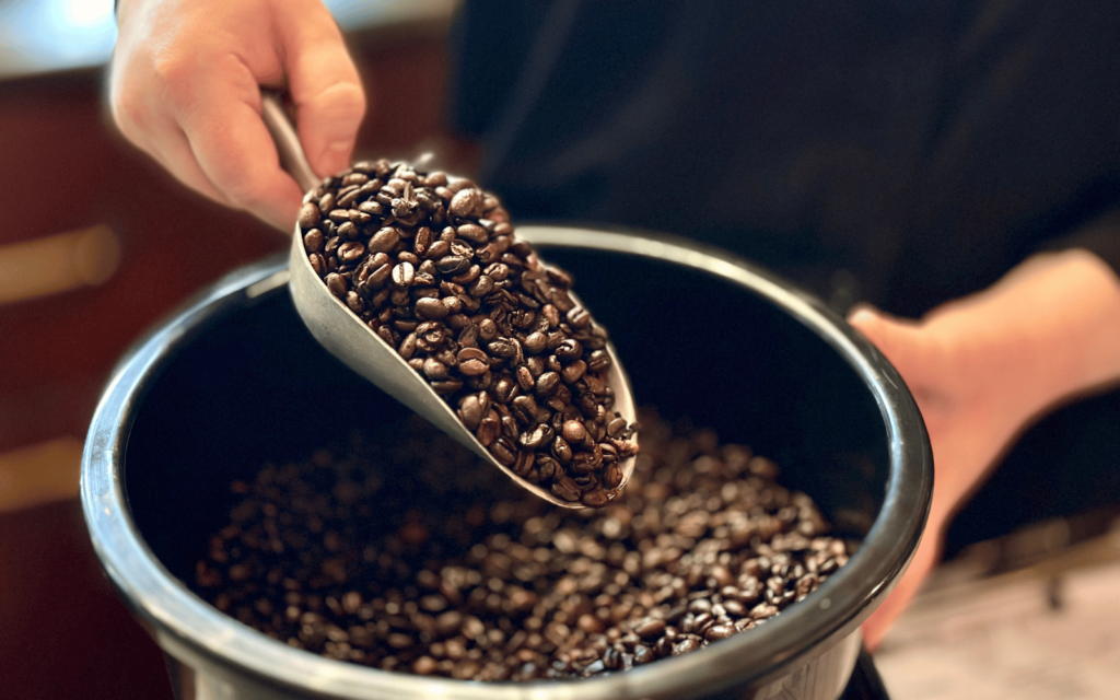 En svart hink med kaffebönor. En hand håller i en skopa fylld med kaffebönor.