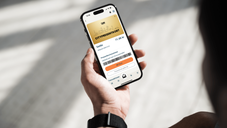 En hand som håller i en mobil. På skärmen ser man ett guldigt kort där det står citypresentkort med svart text på. Ovanför texten ser man siluetten av en stadskärna utgjord av en svart linje. Ovanför siluetten är en svart logga med texten hbg city.
