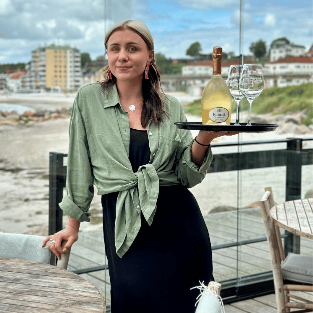 En tjej står med en bricka med en flaska champagne och två champagneglas. I bakgrunden ser man en uteservering och Helsingborgs strand.