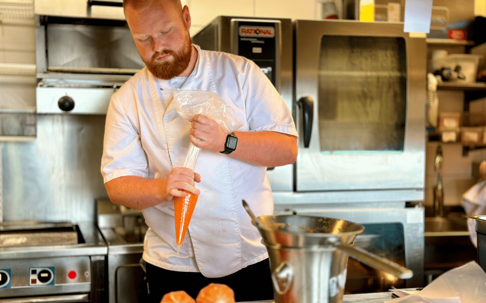 En man i kockkläder står i ett kök och knyter en spritspåse med morotspuré.