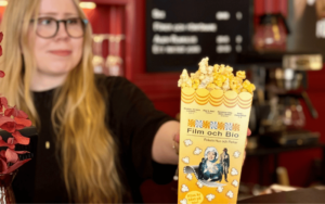 En tjej som står bakom disken i en biograf och lämnar över en burk med popcorn