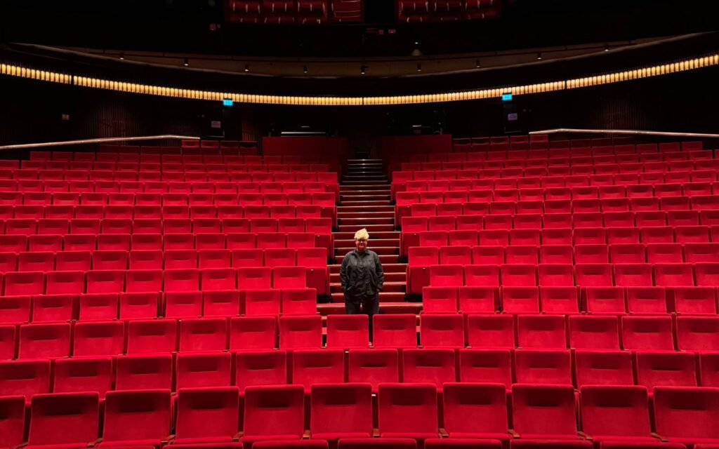 En stor teatersalong med röda stolar och en kvinna som står i svart mitt i bland dom.