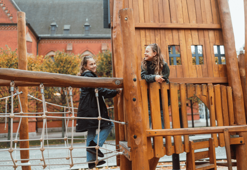 Två flickor leker på en lekplats. I bakgrunden ser man en kyrka och träd med höstlöv.