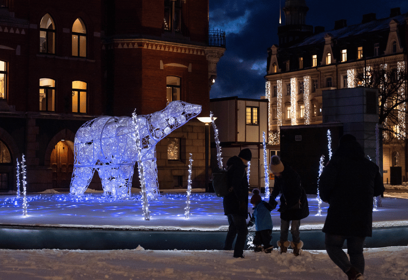 En ljusinstallation som föreställer en isbjörn och istappar. Det är mörkt och det är snö på marken. Framför isbjörnen står tre vuxna och ett barn och tittar på vinterljusen.