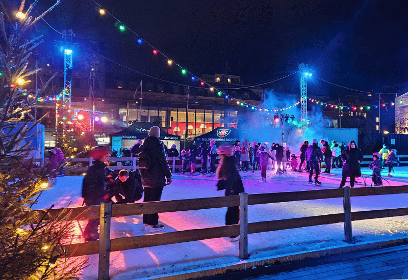 En isrink på ett torg med folk som åker skridskor. Över isbanan hänger lampor i olika färger och strålkastare lyser med ett rosa ljus över isbanan- Det är n rökmaskin som skapar rök på ena hörnet av isbanan. I bakgrunden syns matvagnar.