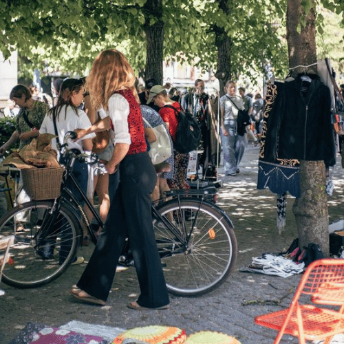 Kvinna leder en cykel på ett torg med loppisstånd i bakgrunden.