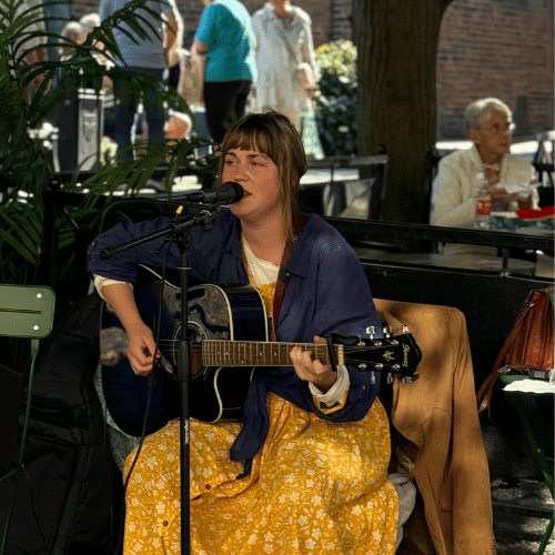 Kvinna spelar guitarr och sjunger i en mikrofon på ett torg.