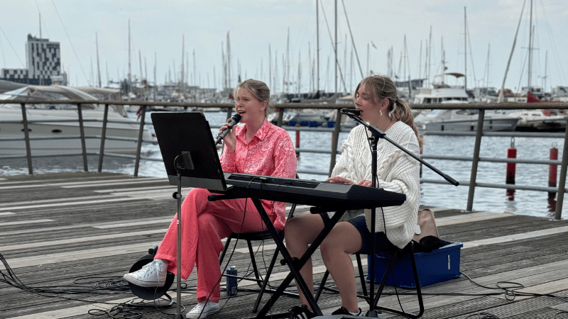 Två tjejer sitter på stolar på en brygga. Tjejen till vänster sjunger i mikrofon och tjejen till höger spelar keyboard. I bakgrunden ser man en hamn med båtar.