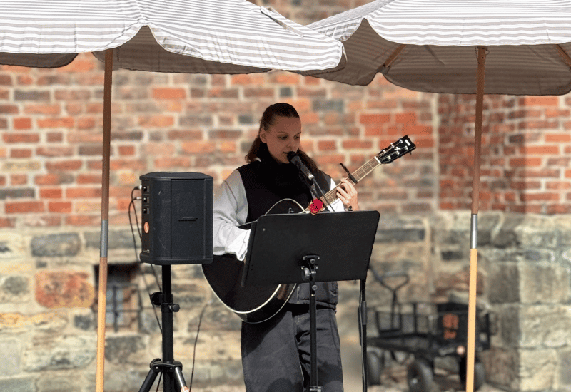 En tjej spelar gitarr och sjunger utomhus. Hon står under två randiga parasoll och framför henne är en högtalare. I bakgrunden ser man en tegelvägg.