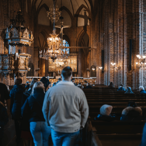 Inuti en gammal kyrka gjord av tegel, det sitter folk på bänkarna och det går folk in mot altaret.