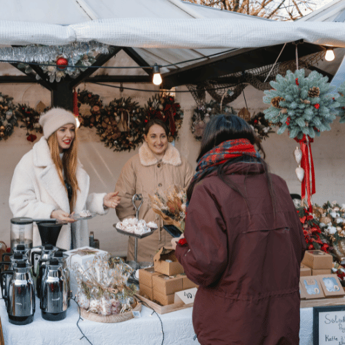 Två kvinnor säljer kransar i ett marknadsstånd och en kvinna står framför och handlar.