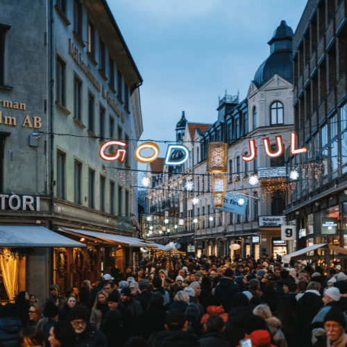 En gågata som är fullproppad med människor under vintertid. Ovanför hänger färgglada bokstäver som lyser, där det står god jul