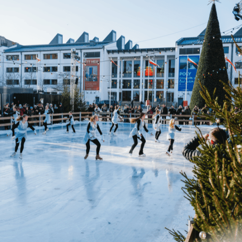 En isbana med barn som åker skridskor. Bakom isbanan står en stor gran som lyser.