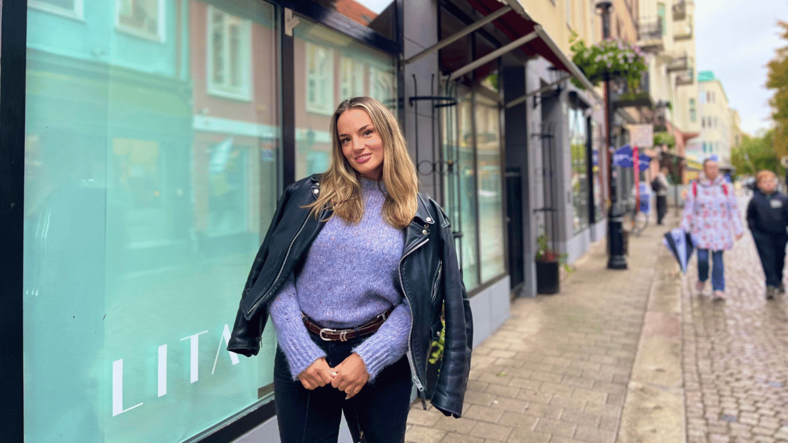 En kvinna i lila, stickad tröja, skinnjacka och svarta byxor står framför en butik på en gågata. På skyltfönstret står det LITA.