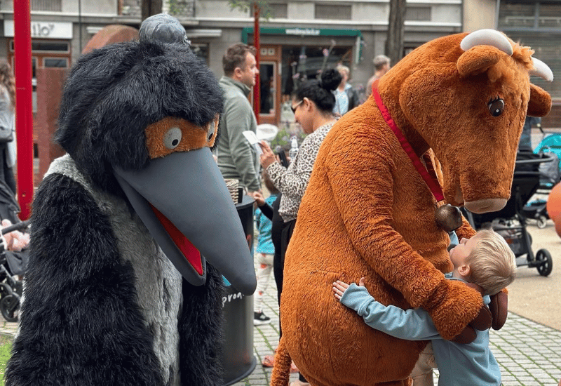 Två maskottar som liknar bok-figurerna Mamma Mu och Kråkan, dom kramar barn på ett torg
