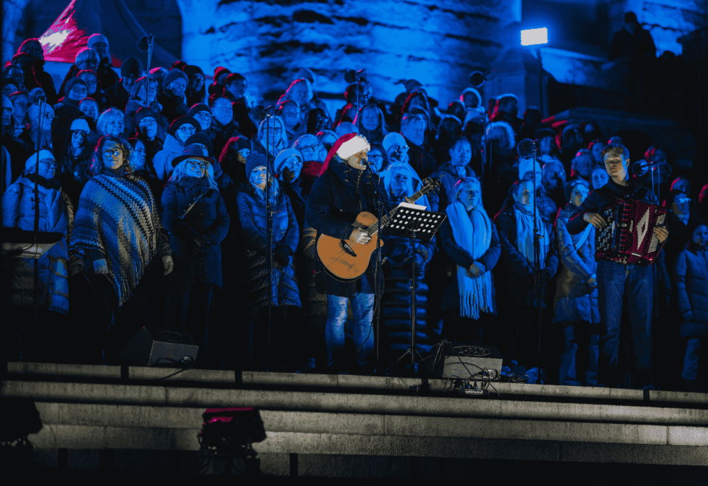 Julshow-framträdande utomhus på trappor. Det står en hel kör och framför en man med tomteluva och gitarr.