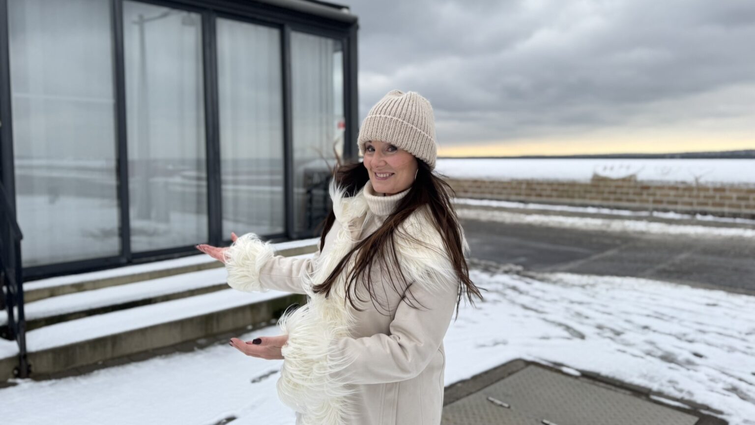 En svarthårig kvinna med beige mössa och beige pälsjacka står framför en glasbyggnad vid havet. Hon ler och pekar inbjudande mot byggnaden.