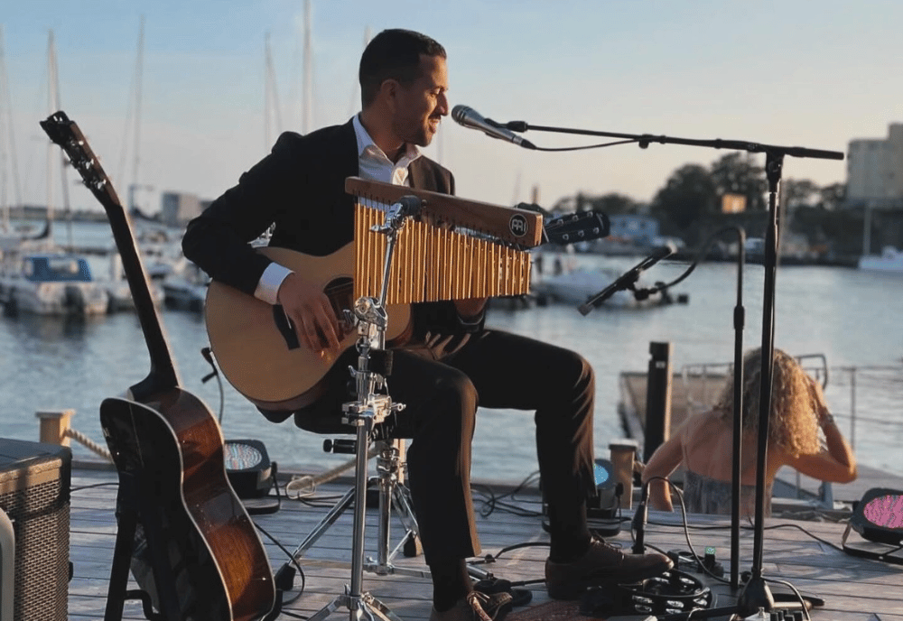 En man sitter på en stol och spelar gitarr på en brygga i en hamn. Runt sig har han flera olika instrument.