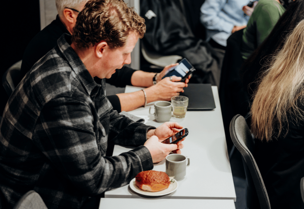 Två män sitter och tittar på sina mobiltelefoner vid ett bord. På bordet står kaffekoppar och frallor.