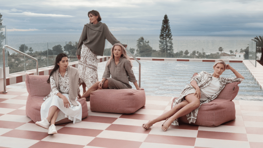 Fyra kvinnor klädda i jordnära färger sitter och står framför en pool.
