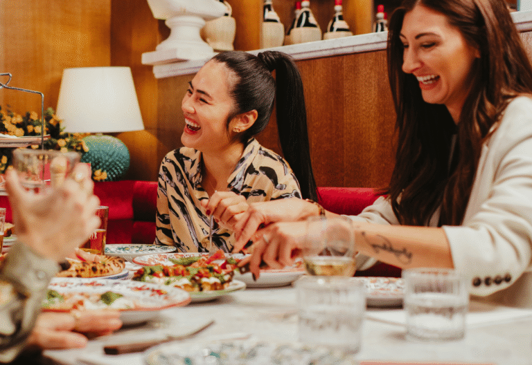 Två kvinnor sitter vid ett bord på en restaurang och skrattar. På bordet står tallrikar och glas.