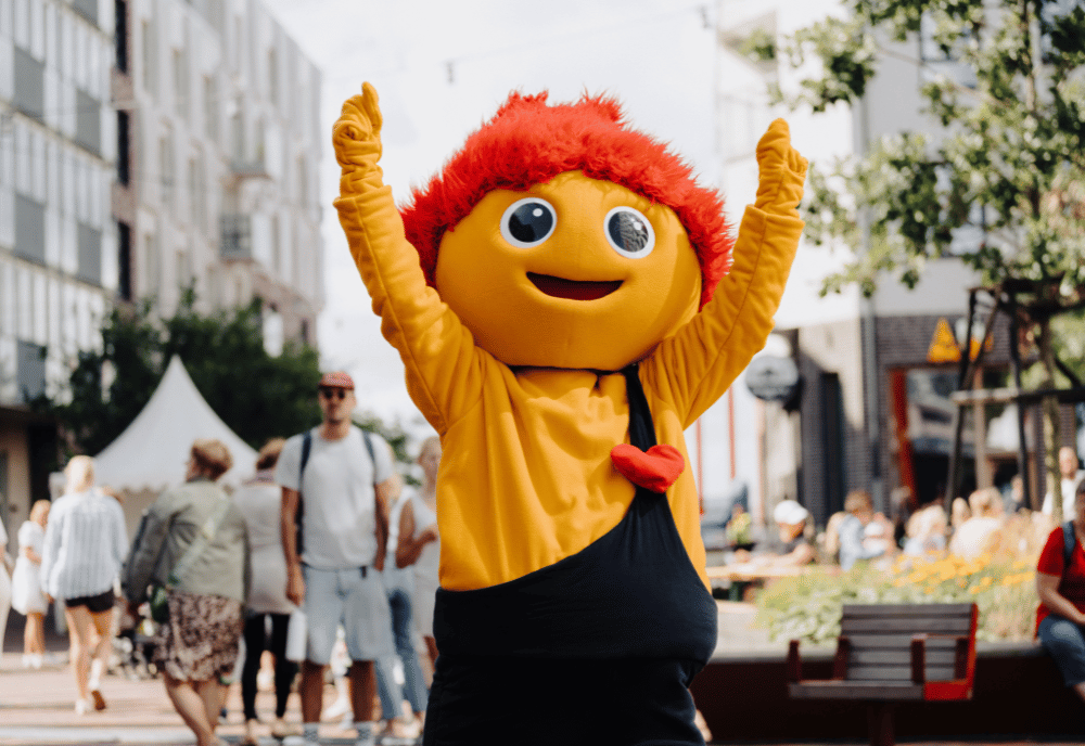 En orange maskor med rött hår och hängslebyxor sträcker upp händerna i luften.