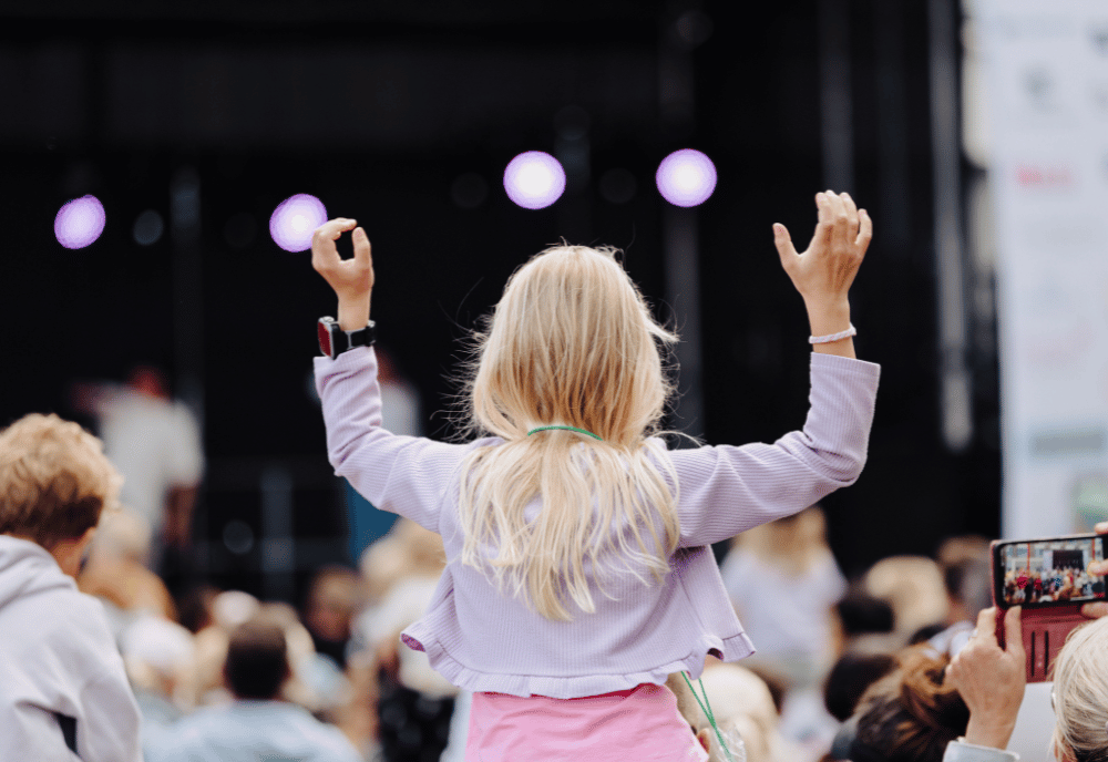 En flicka i lila tröja sträcker upp händerna i luften och tittar mot en scen.