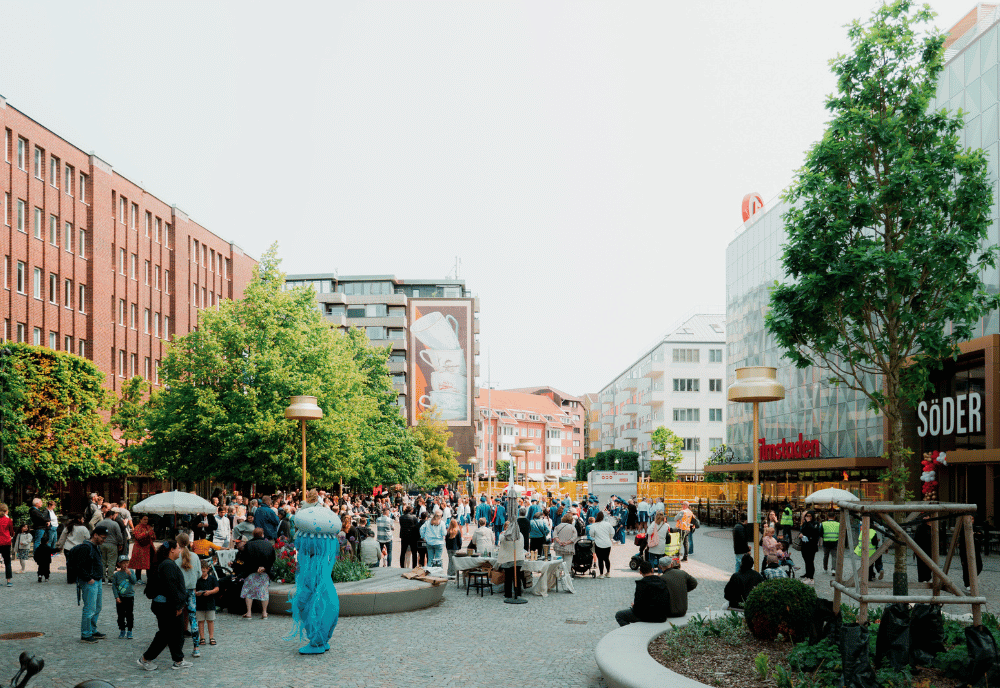 Ett torg med folk och en blå maskot i form av en manet. På torget ser man träd och runt om lägenhetshus och en galleria.