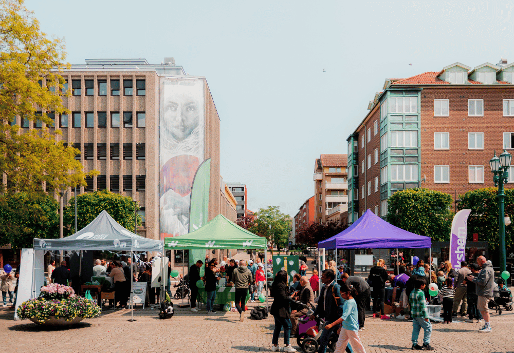 Ett torg med tält och stånd. Runt tälten ser man folk. Runt torget ser man träd och lägenhetshus.
