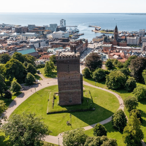 En drönar bild öve en stadsmiljö, ett stort torn i mitten av en gräsmatta och utsikt över stad och hav