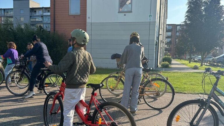Barn i olika åldrar som gåller i cyklar på en gata med grönområde och hus i bakgrunden